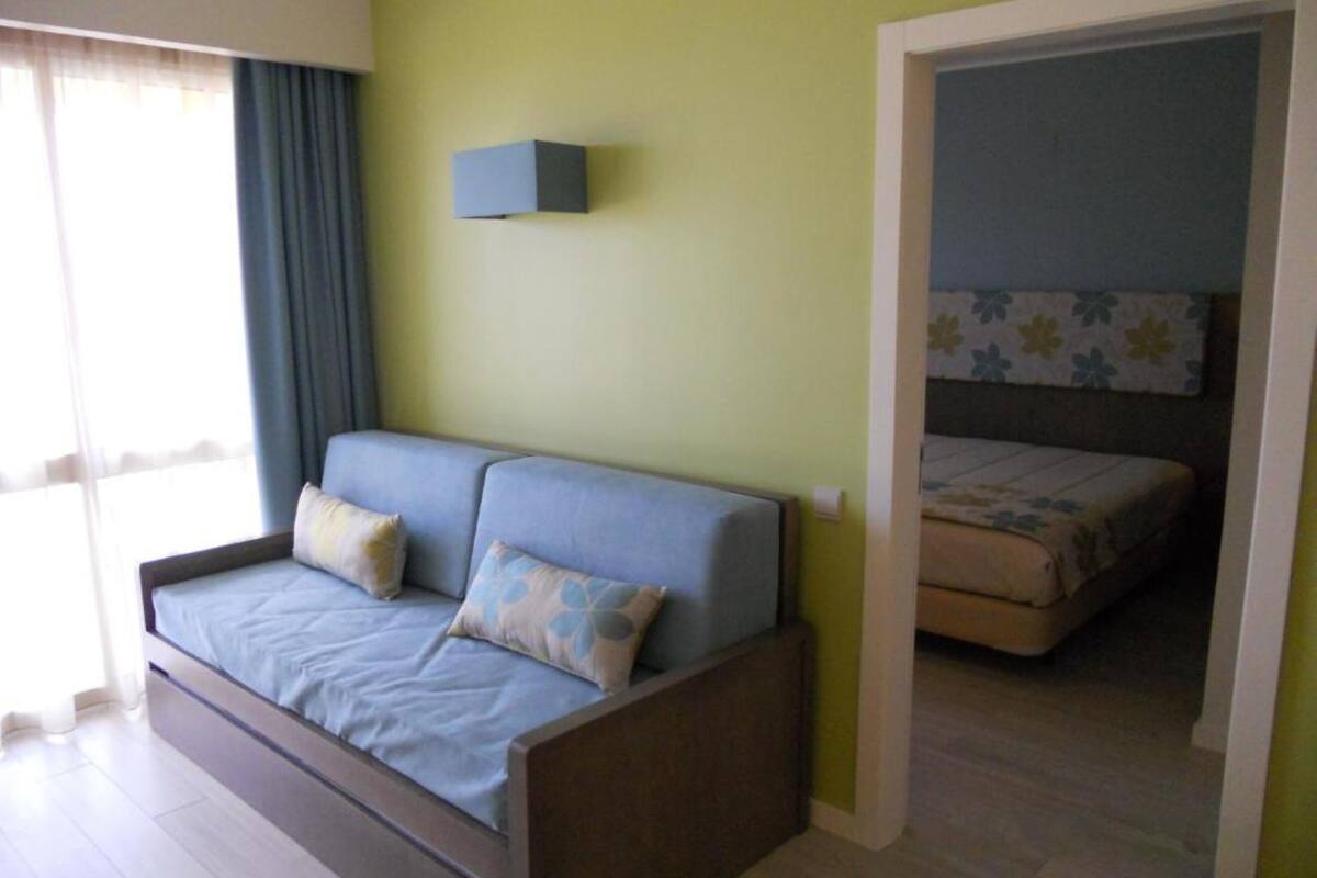 Een kamer met een slaapbank, een klein nachtkastje, een wandlamp en een bed met een bloemenbedekking.