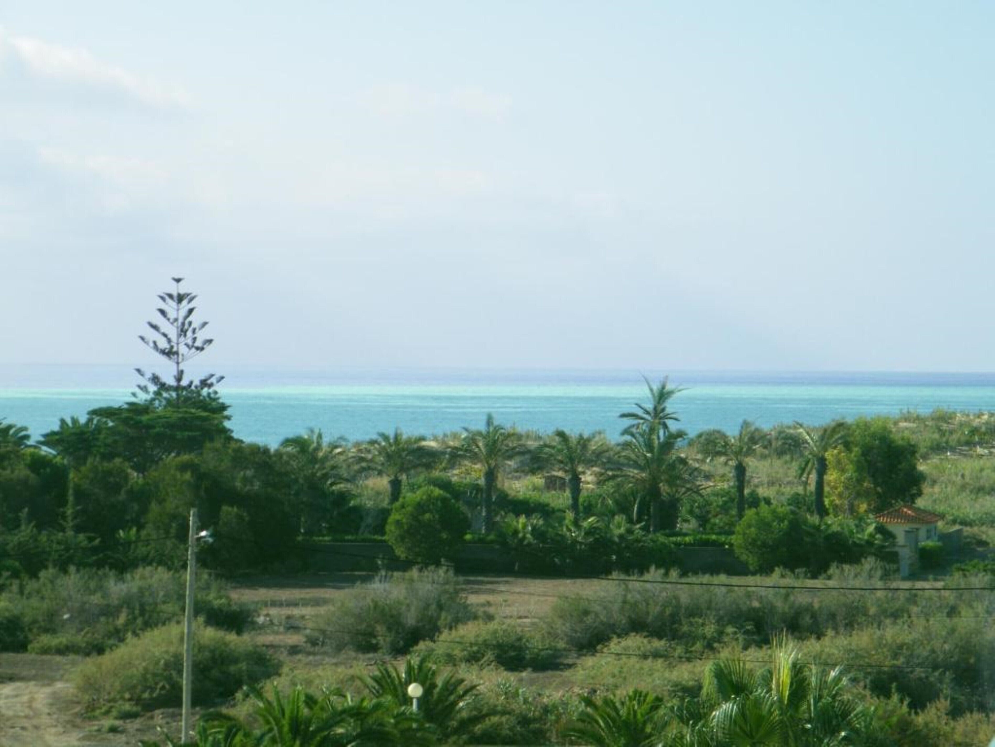 A coastal landscape with a clear view of the sea, dense greenery, and a few scattered buildings.