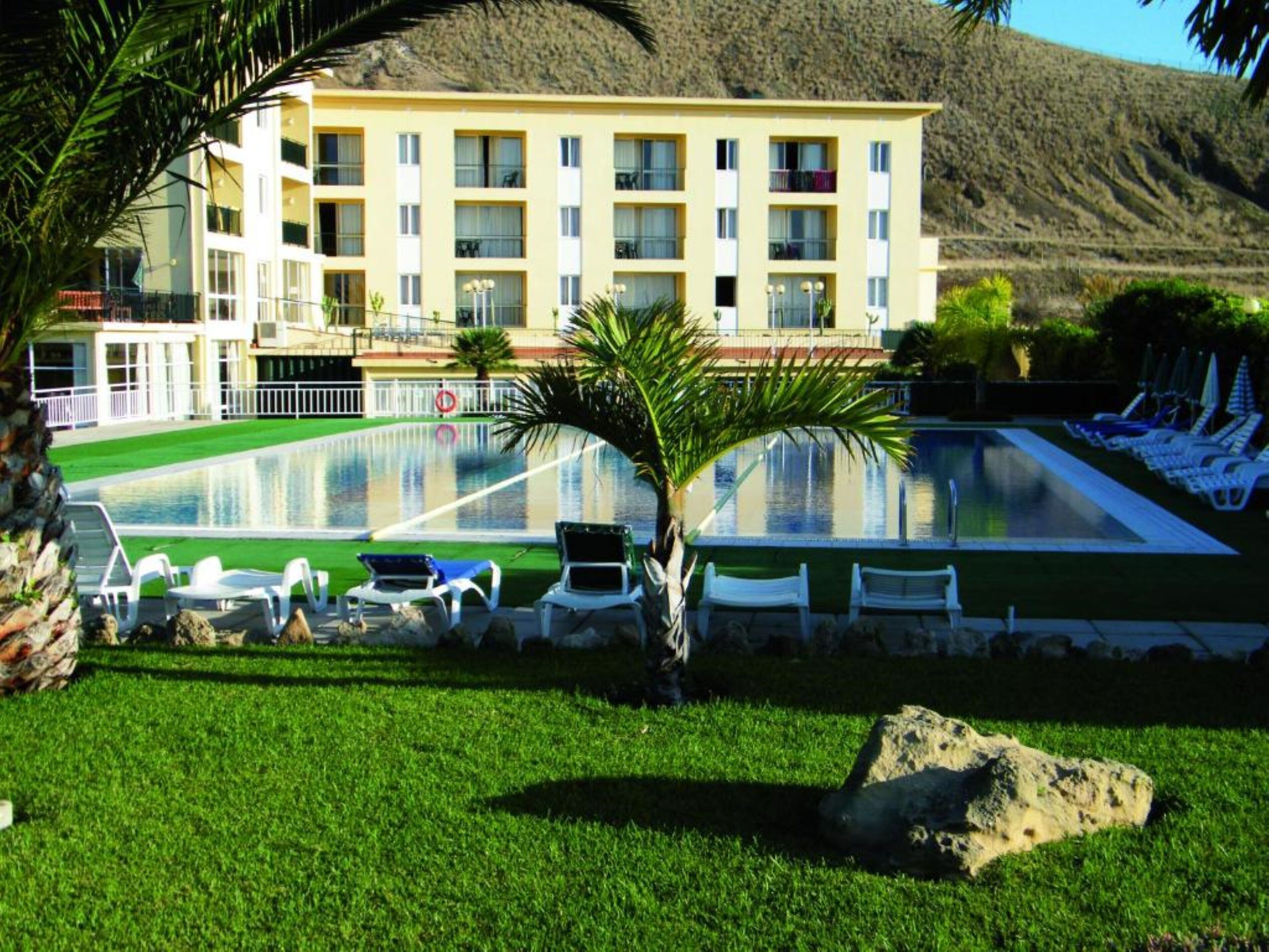 Un hotel con piscina, sillones de descanso y montañas al fondo.