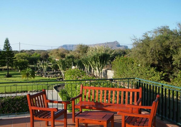 A terrace with a wooden bench and table, surrounded by a green railing, overlooking a landscaped garden and distant mountains.