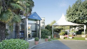 Entrada de un hotel con un letrero azul, puerta de vidrio y una zona cubierta con toldo que tiene mesas y sillas.