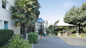 Entrada de un hotel con un área de estar al aire libre techada, una palmera y un edificio con toldo azul.