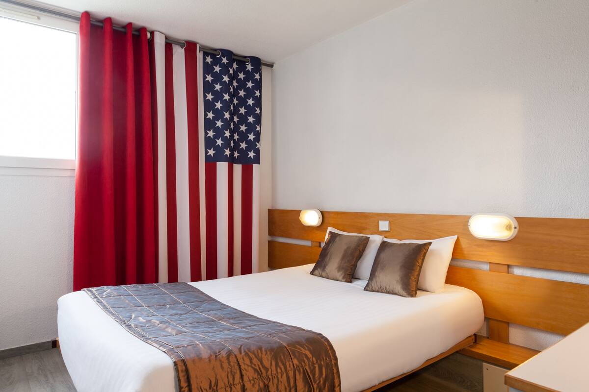 Een hotelkamer met een bed, een hoofdeinde met verlichting, een raam met rode gordijnen en een Amerikaanse vlag.