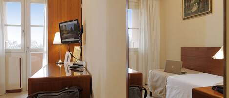 Una habitación de hotel con un escritorio de madera, una computadora portátil, una cama con edredón blanco, un cuadro en la pared y vistas al exterior a través de la ventana.