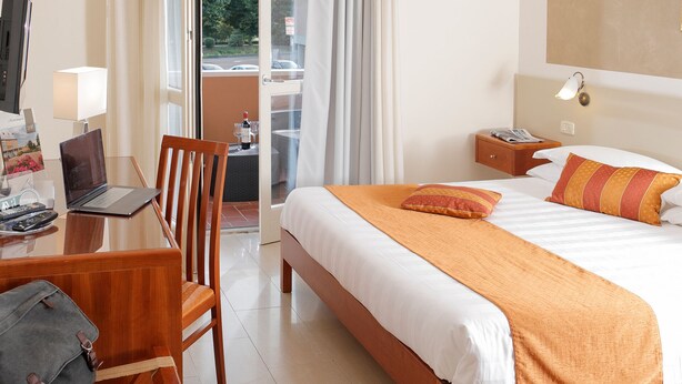 A hotel room with a bed, a desk with a laptop, a chair, a nightstand, and a view through the window. - Hotel Michelangelo (Terranuova Bracciolini)