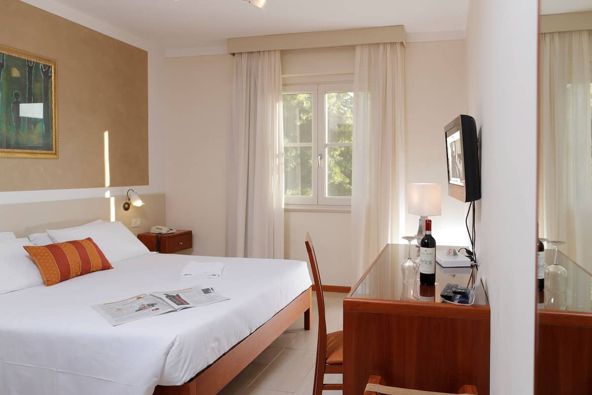 Una camera d'albergo con un letto, una scrivania con una sedia, una televisione e una finestra con tende.