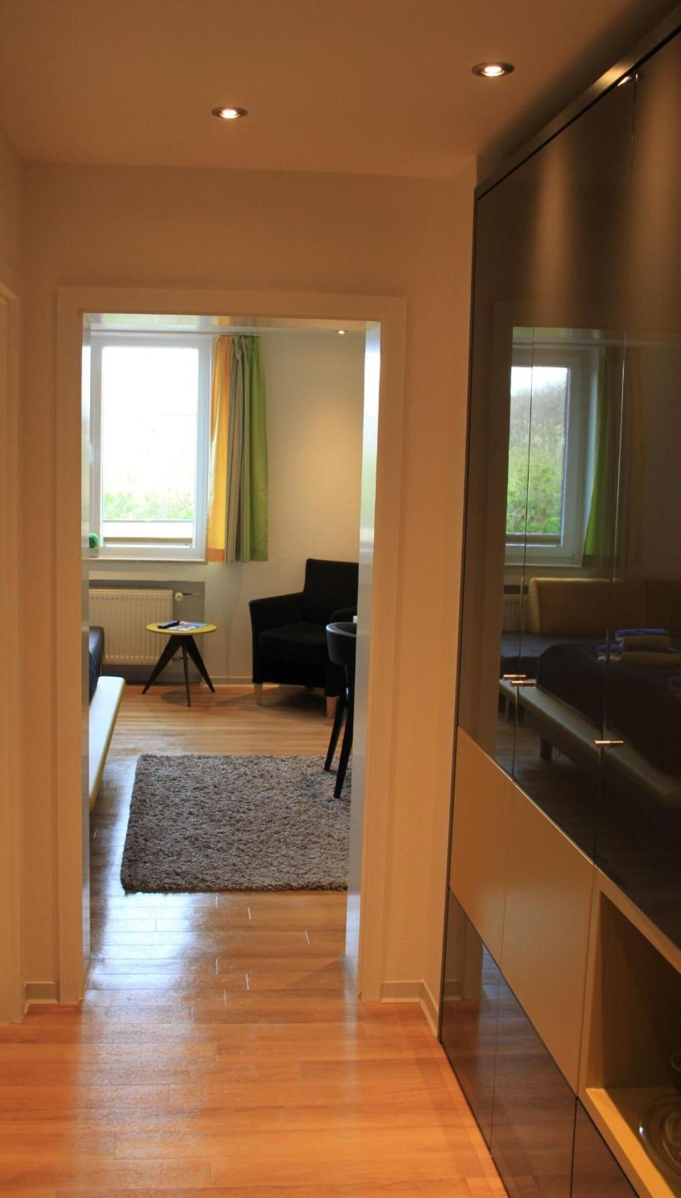 Un interior moderno con piso de madera, un sillón negro, una mesita redonda y vista a un paisaje verde a través de una ventana.