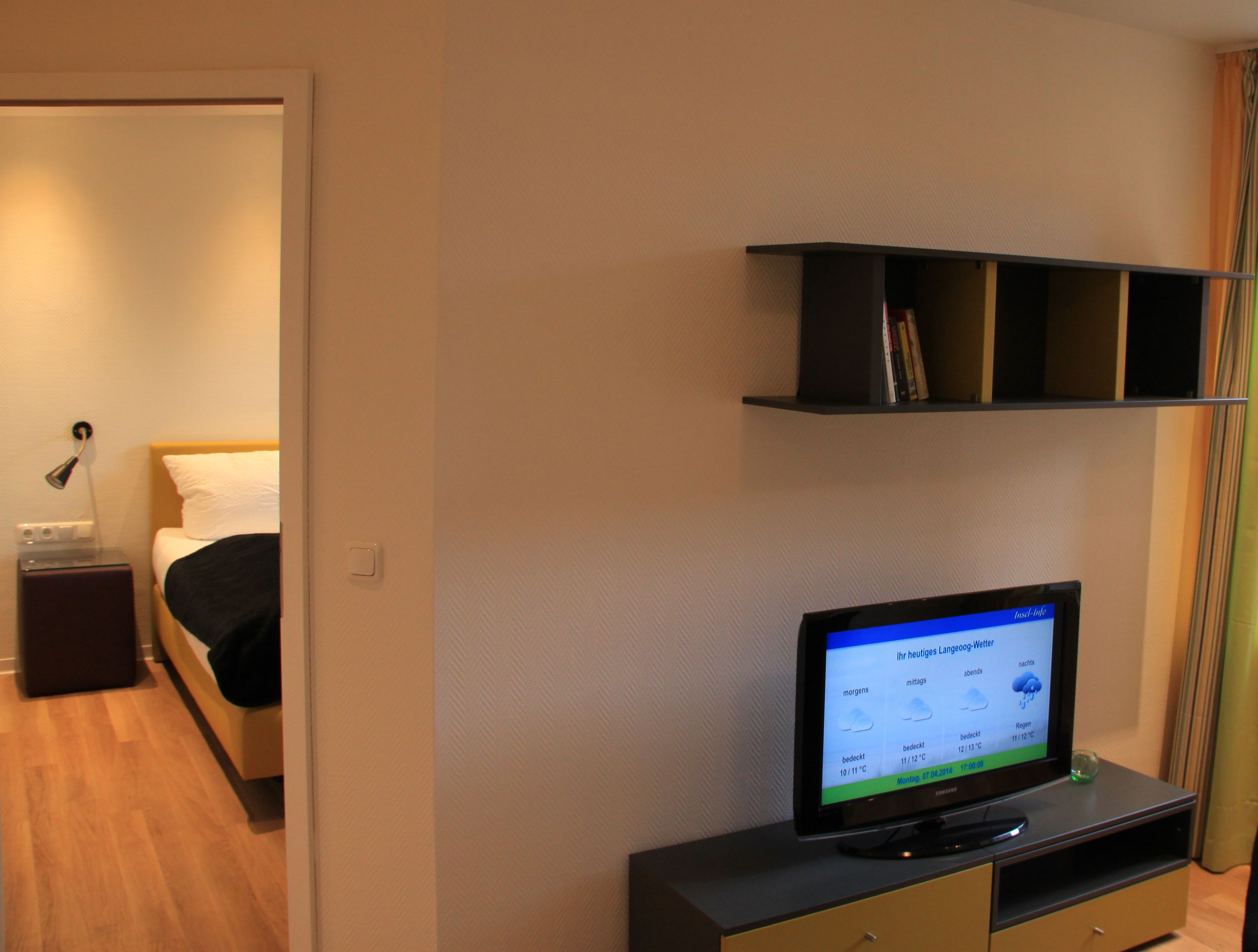 A hotel room with a bed, bedside table, lamp, and a television displaying a weather forecast.