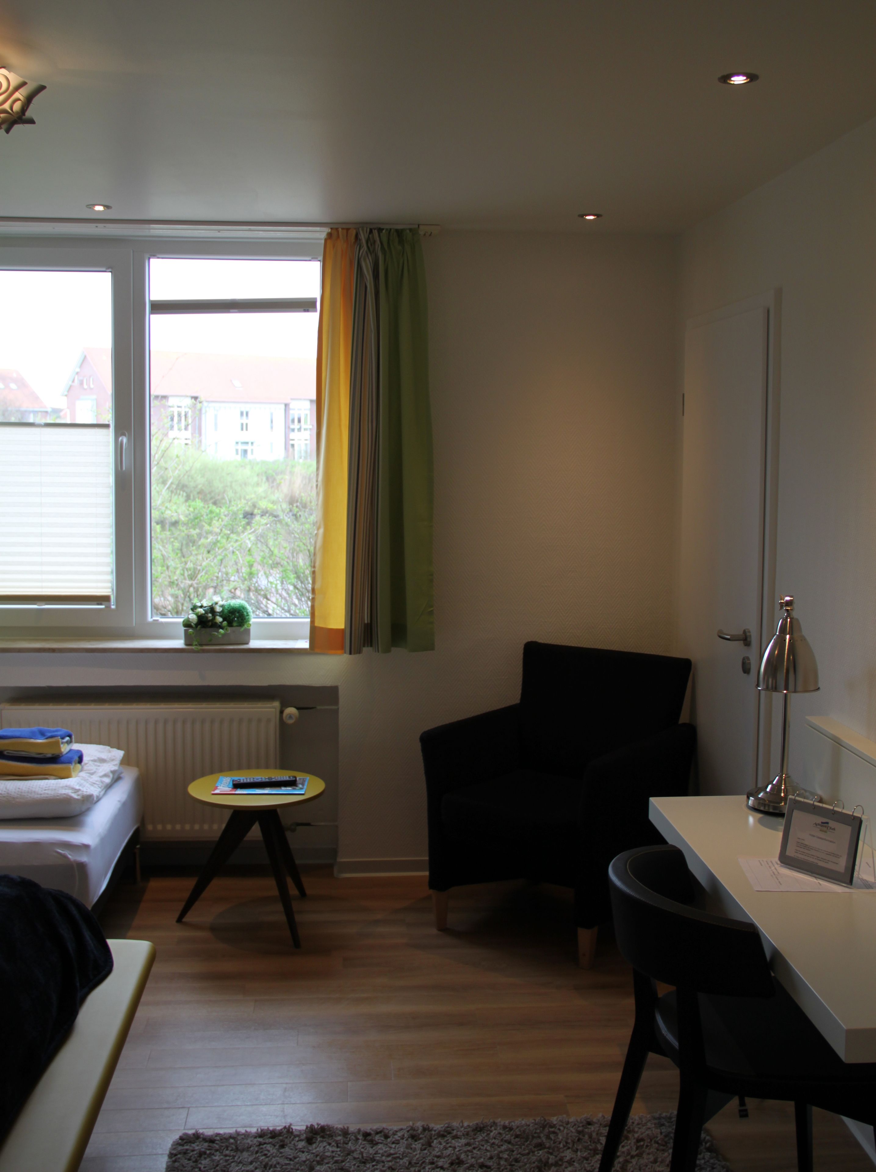 Habitació d'hotel senzilla amb llit, escriptori petit, cadira, radiador i finestra amb cortines.