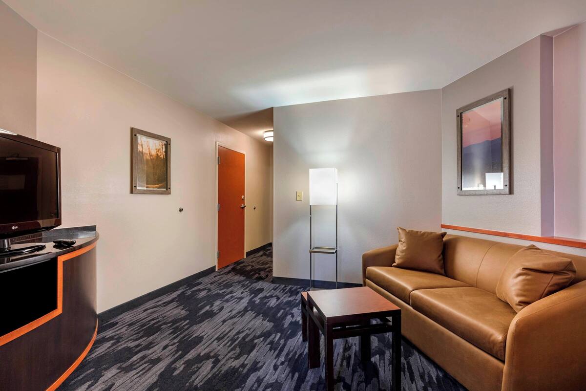 A hotel room with a brown leather sofa, a small wooden table, a TV on a wooden stand, a framed picture on the wall, and a door leading to another room.