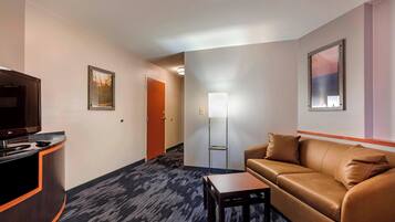 A hotel room with a brown leather sofa, a small wooden table, a TV on a wooden stand, a framed picture on the wall, and a door leading to another room.