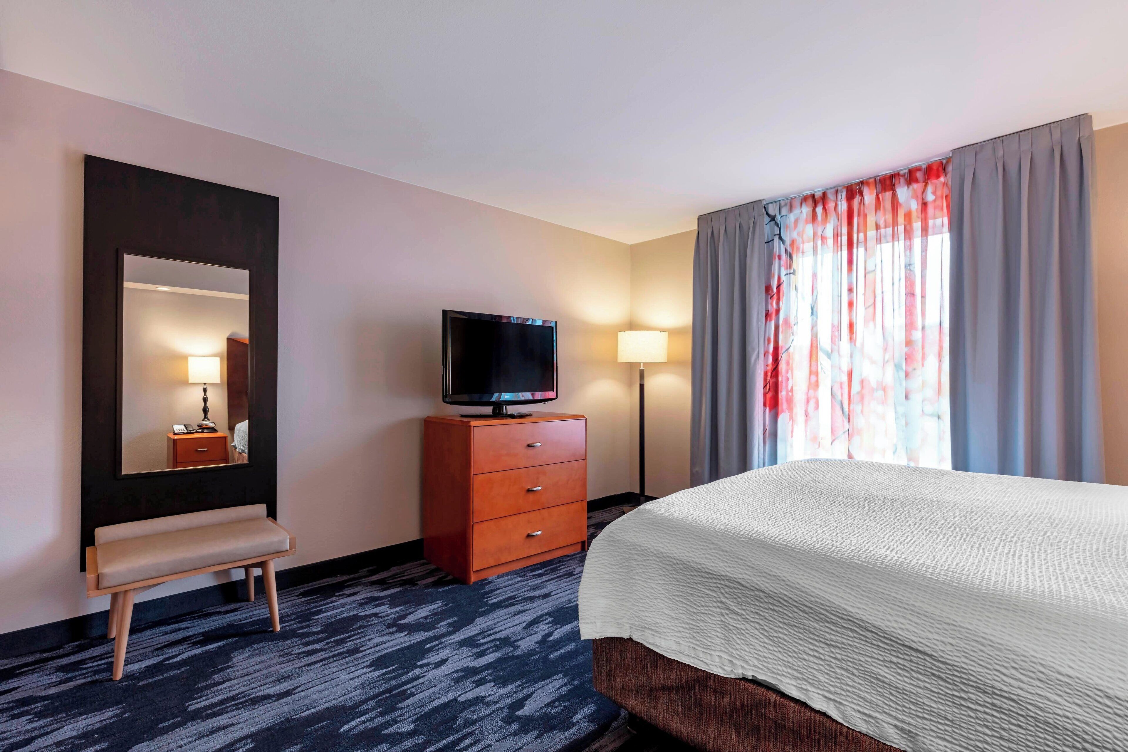 A hotel room with a bed, a television on a wooden dresser, a bench, a mirror, and a window with curtains.