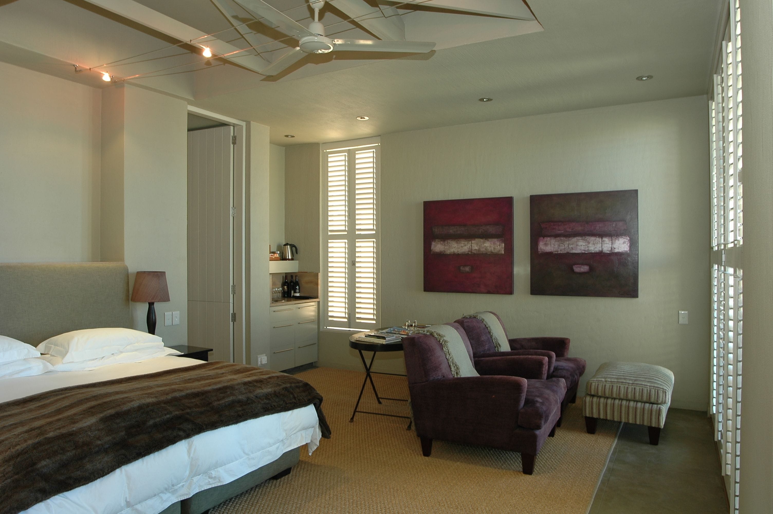 A hotel room with a bed, a desk, a chair, an ottoman, and two abstract paintings.