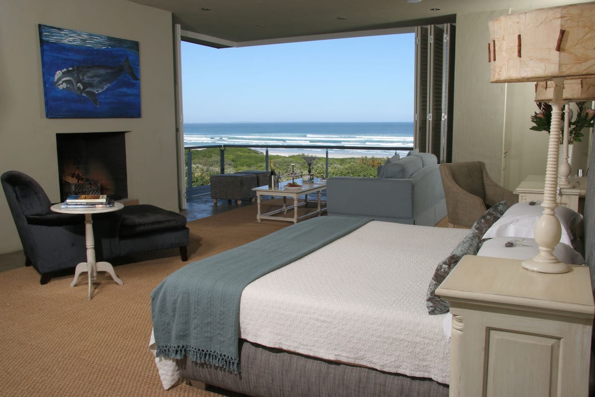 A bedroom with a large bed, a fireplace, a view of the ocean, and a balcony with outdoor seating.