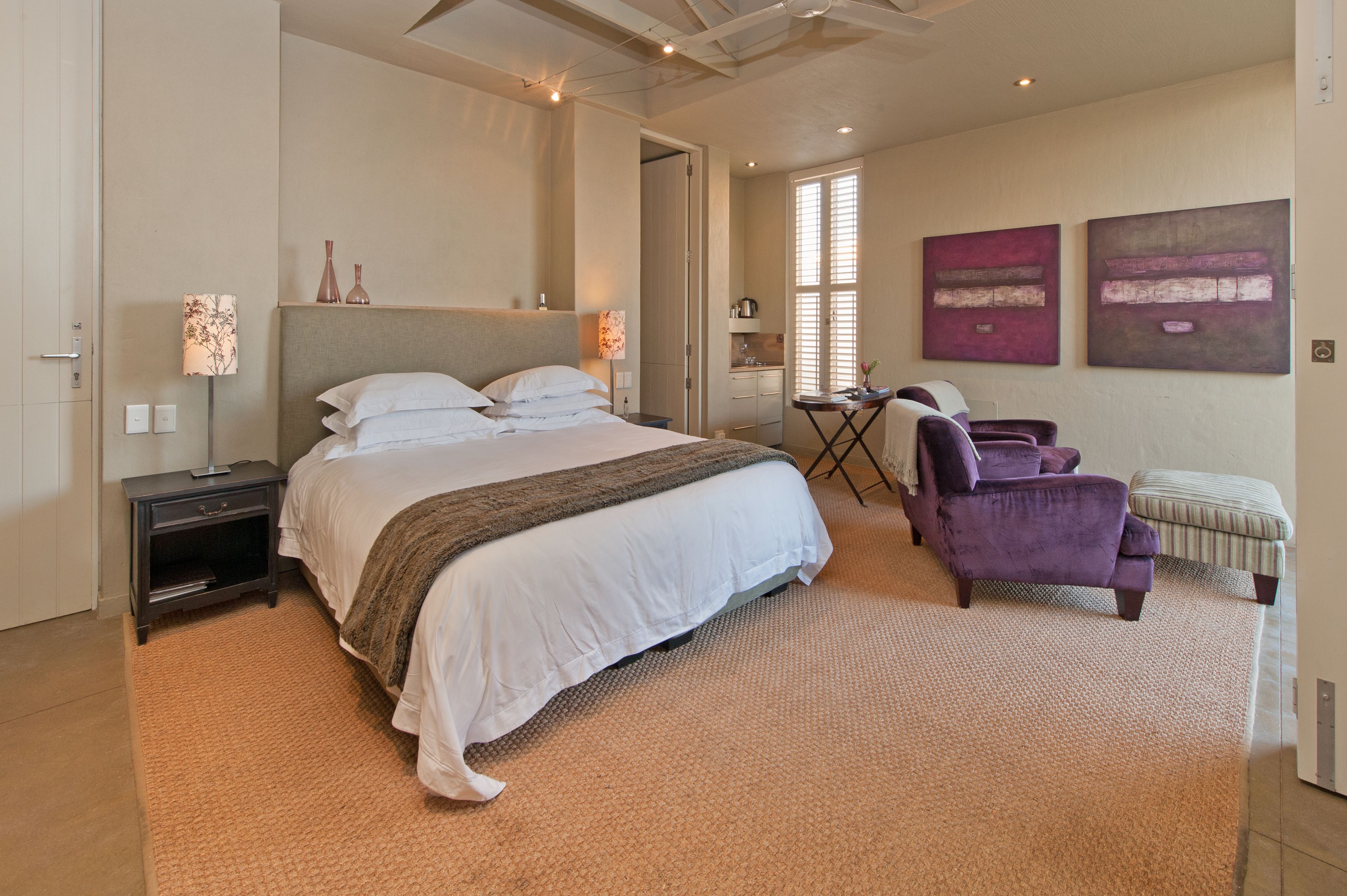 A bedroom with a large bed, a nightstand, a chair, an ottoman, and two artwork pieces on the wall.