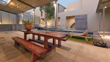 A modern outdoor patio with a wooden table and benches, a pool, and a covered seating area.