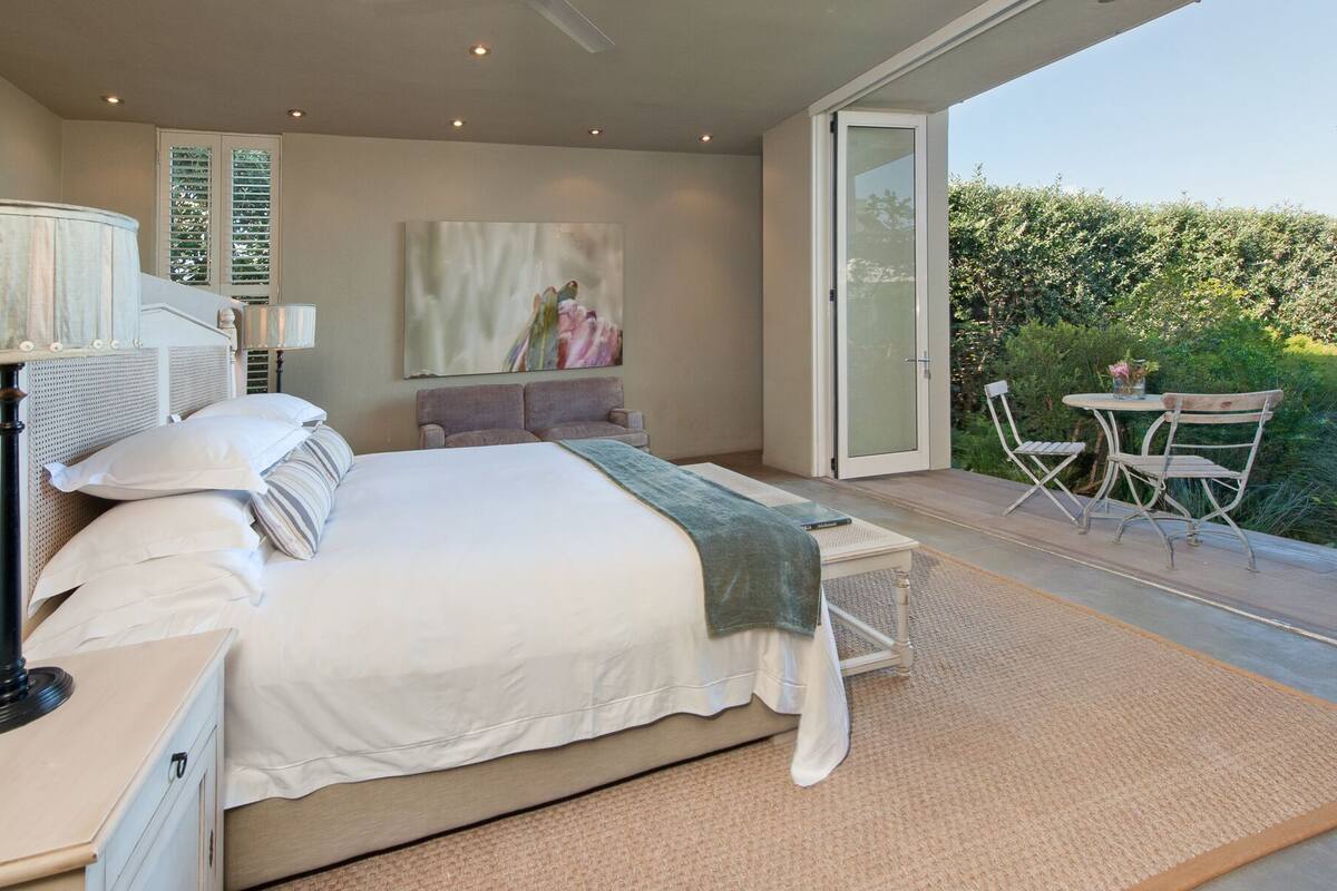 A bedroom with a large bed, a bedside table, a chair, a small table with a vase, and a view of a garden through a sliding door.