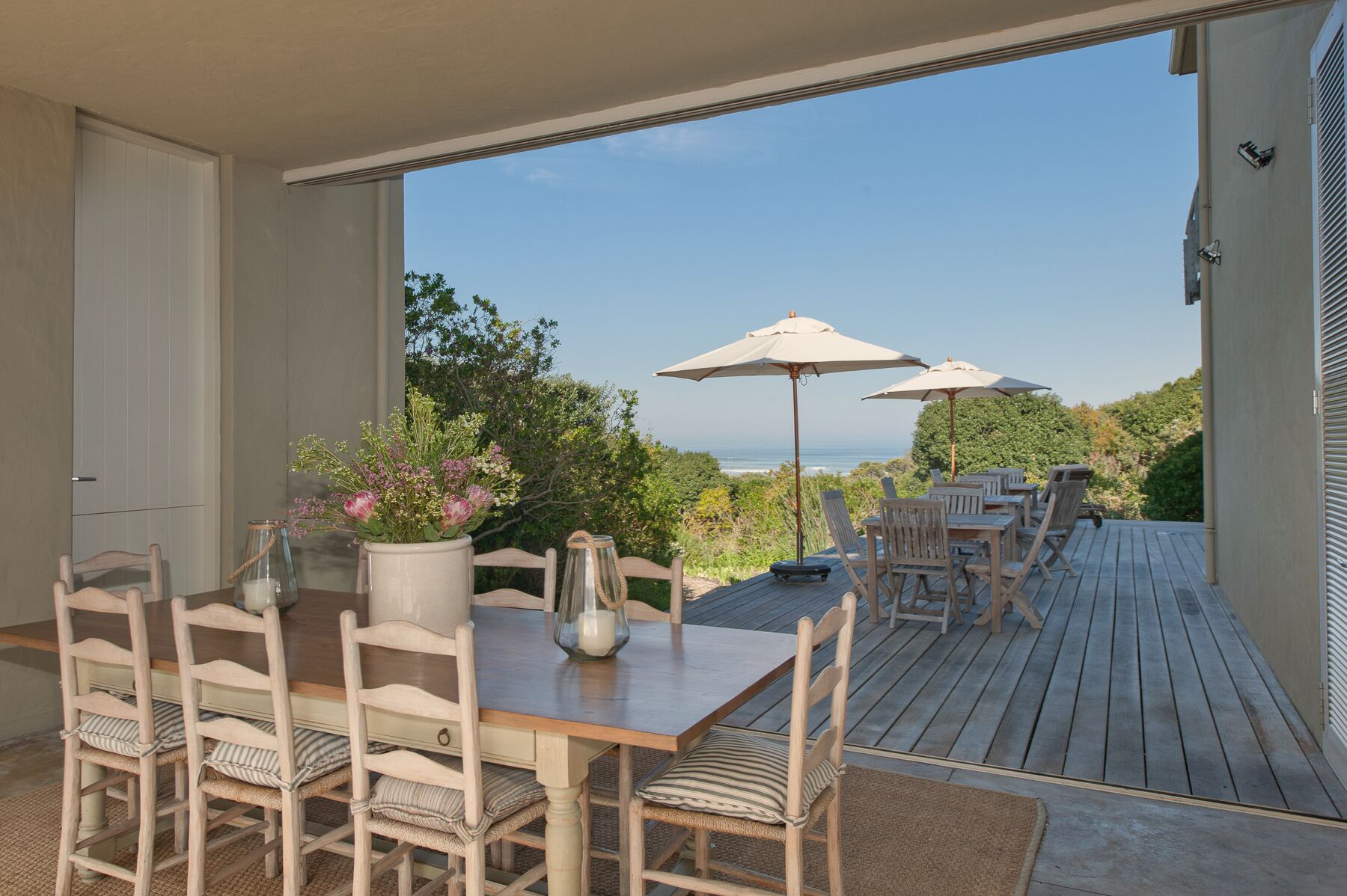 Una terraza de madera con mobiliario exterior, incluyendo una mesa y sillas, y dos sombrillas que dan sombra.