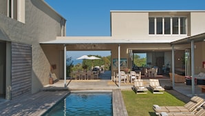 A modern outdoor pool area with lounge chairs, a covered patio, and a view of the backyard.