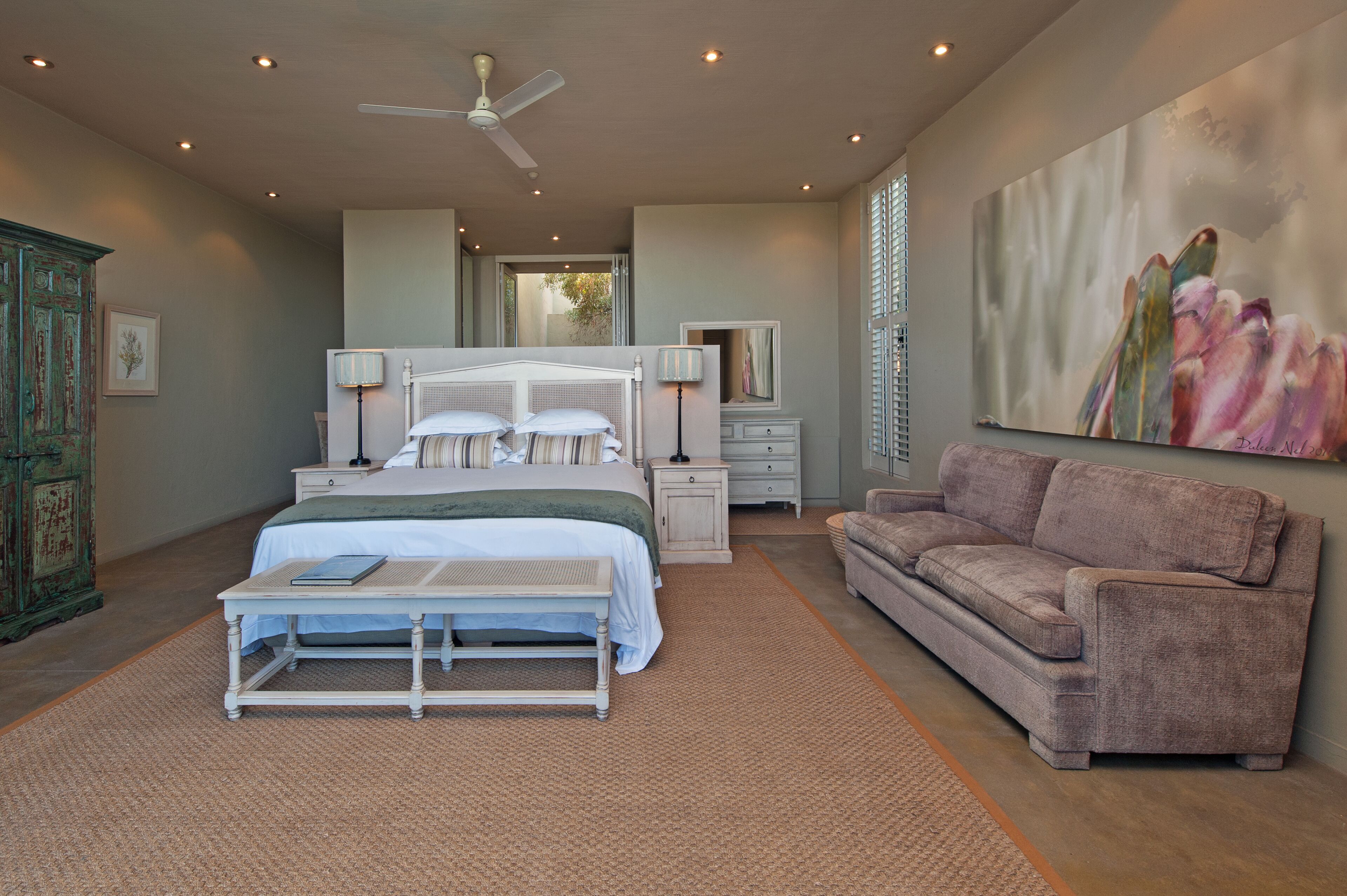 A bedroom with a bed, bedside tables, a bench, a sofa, a ceiling fan, and a large painting on the wall.