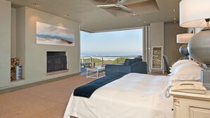 A modern bedroom with a large bed, a fireplace, a view of the ocean, and a balcony with outdoor furniture.