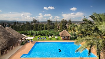 A swimming pool with lounge chairs and umbrellas, surrounded by tropical vegetation and a view of a distant city.