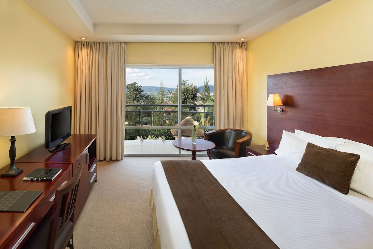 Habitación de hotel con una cama grande, un escritorio con televisor, una silla y vistas al exterior a través de un ventanal amplio.