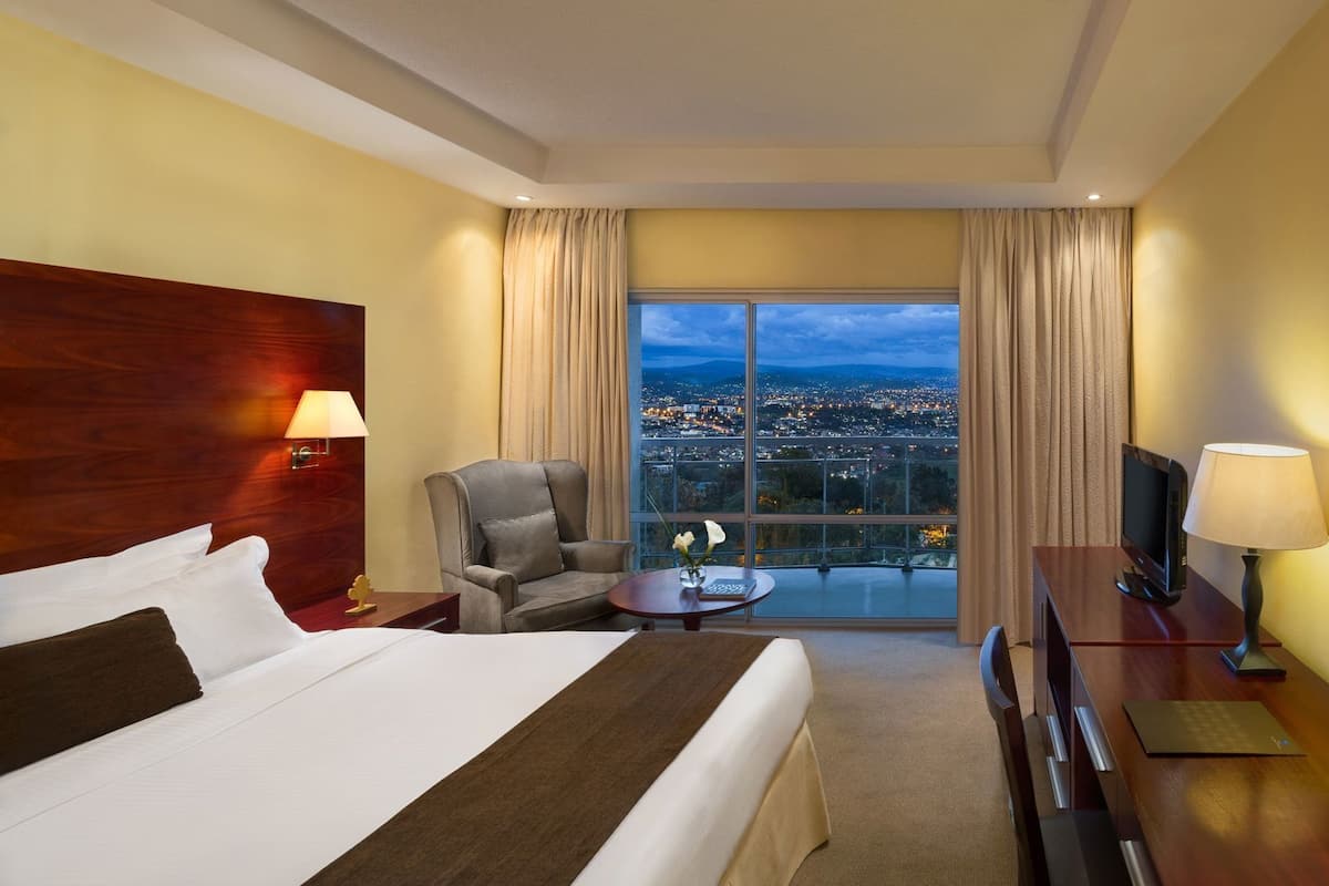 Habitación de hotel con una cama grande, un escritorio, una silla, un televisor y vistas a la ciudad.