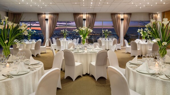 Un salón de banquetes con mesas redondas dispuestas para comer, sillas blancas y grandes centros de mesa florales.