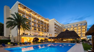 A hotel with multiple balconies, a swimming pool, and a thatched-roof structure.