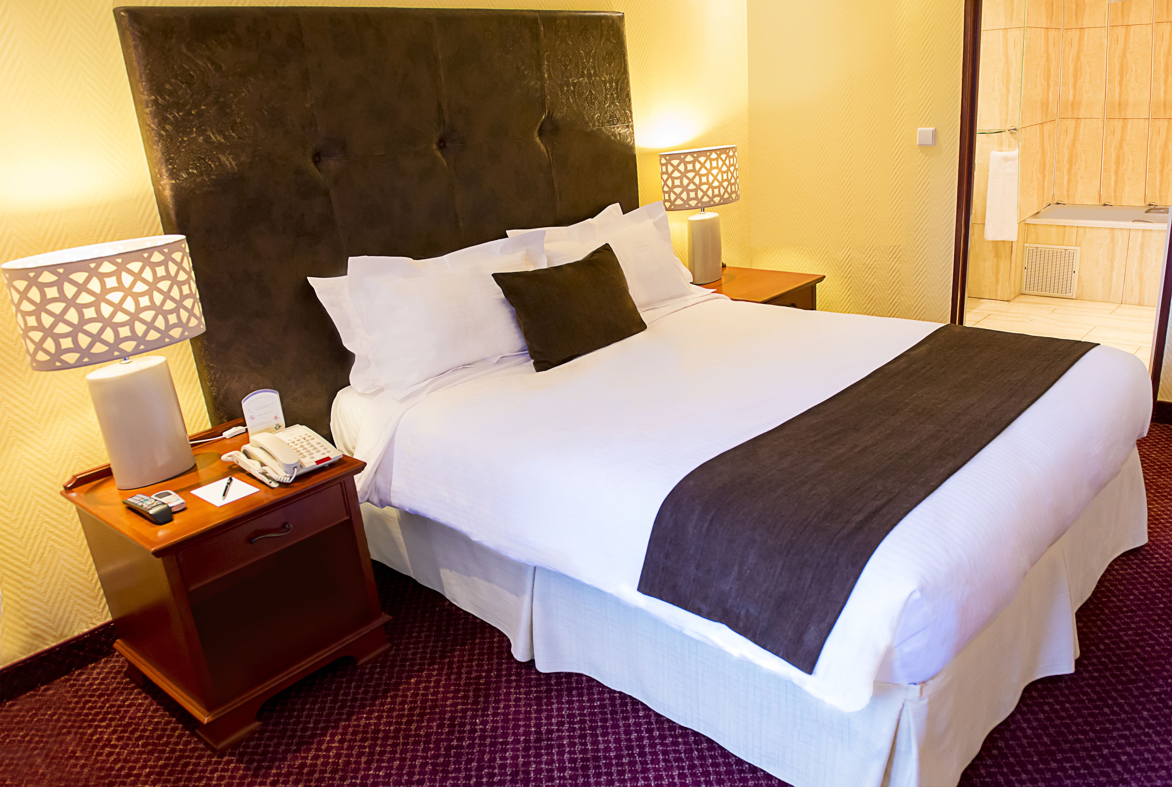 Habitación de hotel con una cama grande, una mesita de noche de madera con un teléfono y una lámpara, y un baño visible a través de una puerta abierta.