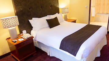 Une chambre d’hôtel avec un grand lit, une table de chevet en bois sur laquelle se trouvent un téléphone et une lampe, et une salle de bain visible à travers une porte ouverte.
