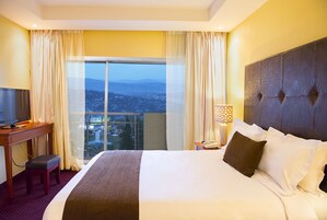 A hotel room with a large bed, a television, a desk, and a view of a cityscape through a large window.