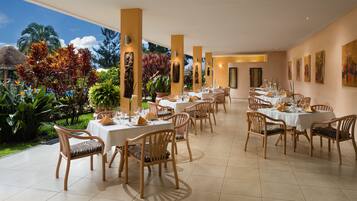 A restaurant with multiple tables and chairs, surrounded by tropical plants and a pool.