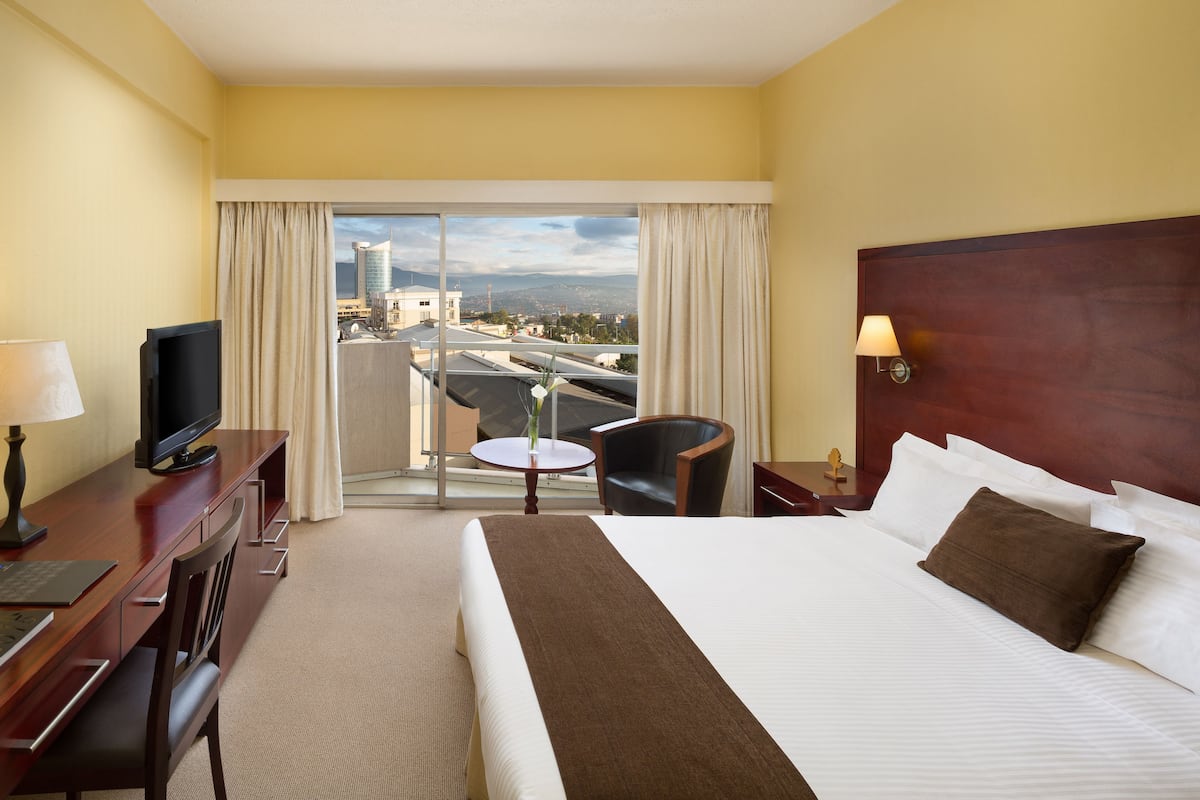 Habitación de hotel con una cama grande, un escritorio con televisor, una silla, una mesita con un jarrón y vistas a la ciudad desde el balcón.