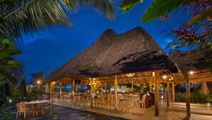 Un área de comedor al aire libre con techo de paja, mesas y sillas de madera, rodeada de plantas tropicales.