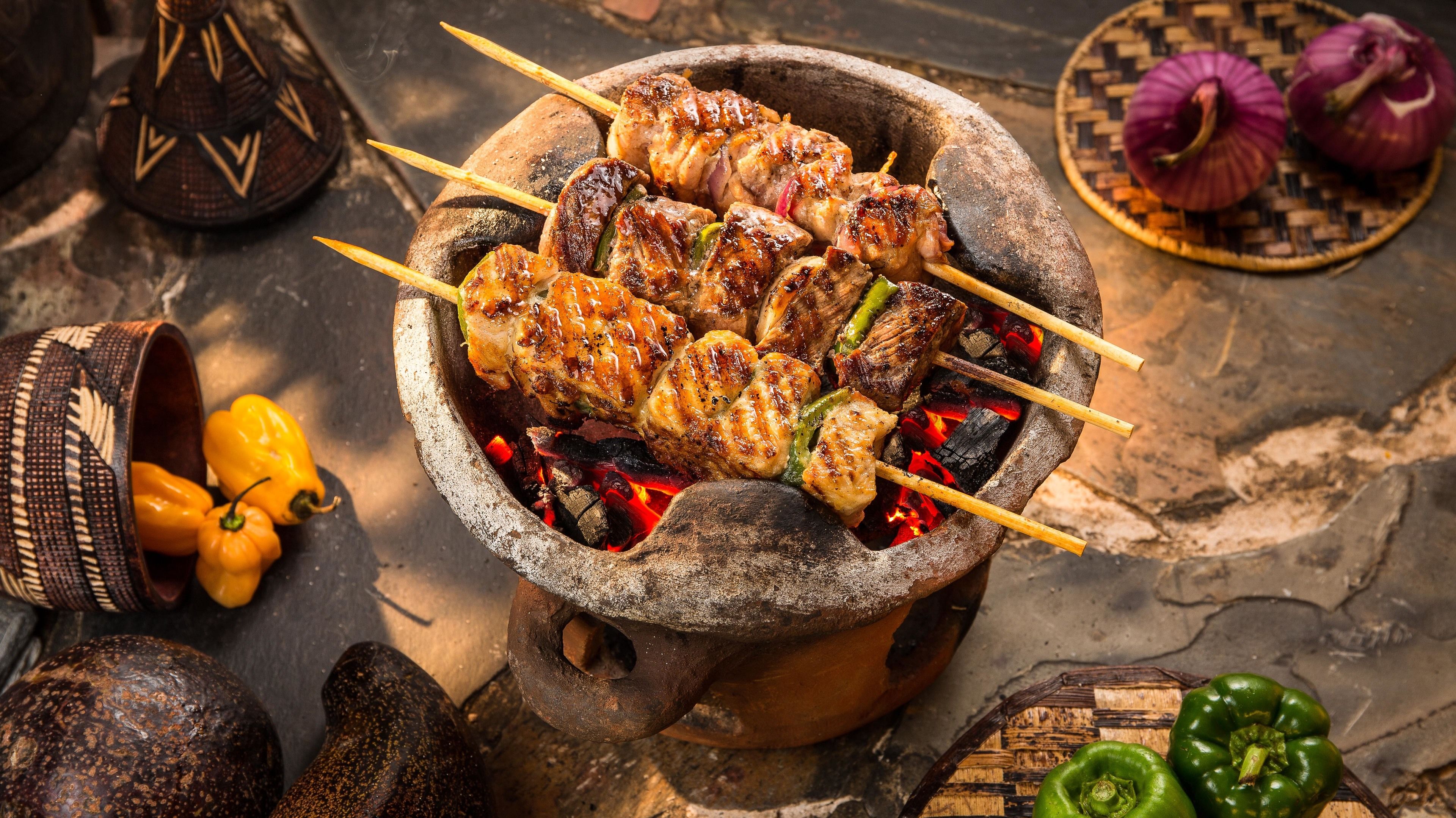 Montaje de cocina tradicional con brochetas de carne y verduras sobre fuego abierto, rodeado de diversos utensilios y alimentos.