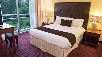 A hotel room with a large bed, two bedside tables, a desk, and a view of a garden through a sliding door.