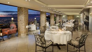 Un área de comedor en la azotea con pilares de piedra, mesas redondas dispuestas para cenar y vistas de la ciudad al anochecer.