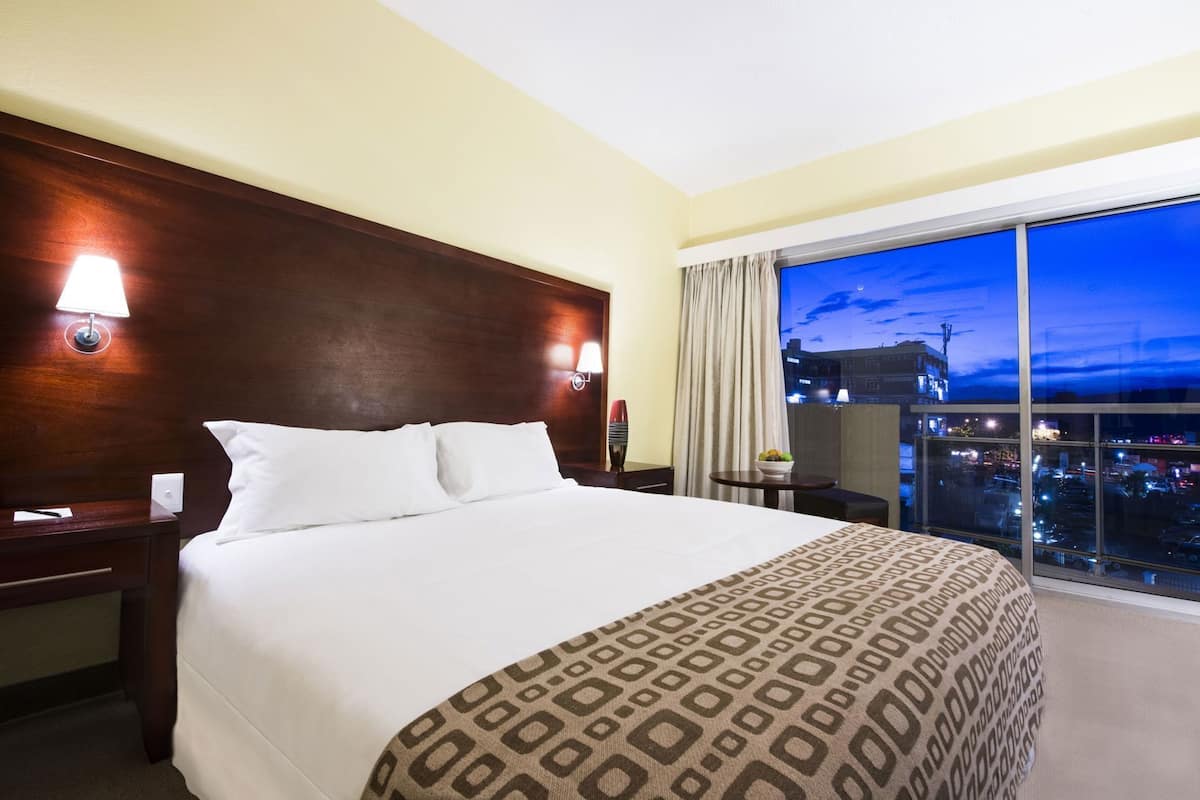 Habitación de hotel con una cama grande, dos mesitas de noche, un ventanal con vista a la ciudad y un balcón.