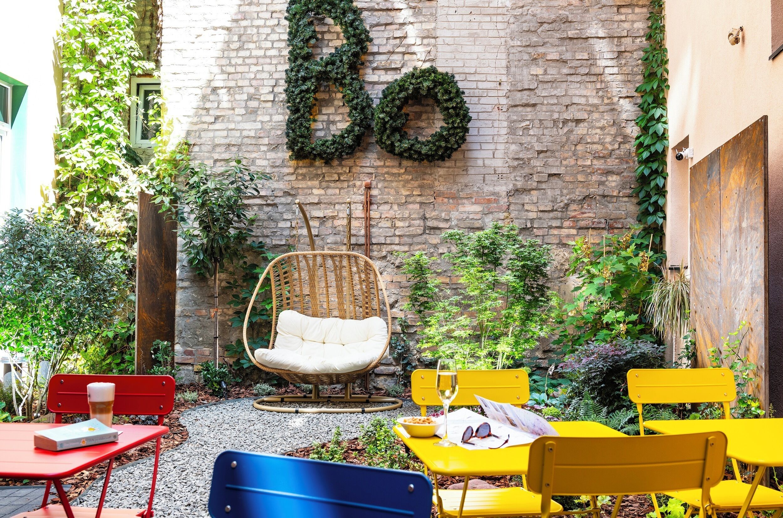 Un patio con pared de ladrillo, una silla de mimbre, sillas amarillas y rojas, y una mesa roja con un libro.