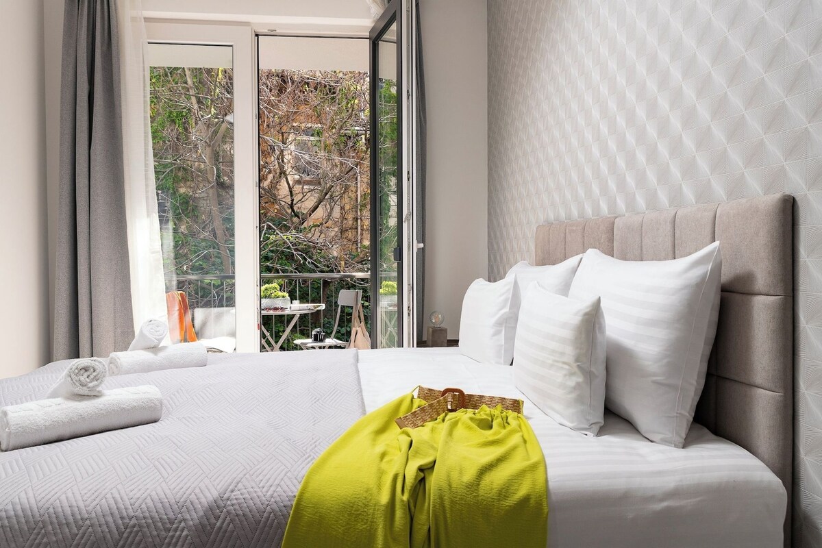 A hotel room with a bed, a window with curtains, a view of trees, and a balcony with a table and chairs.