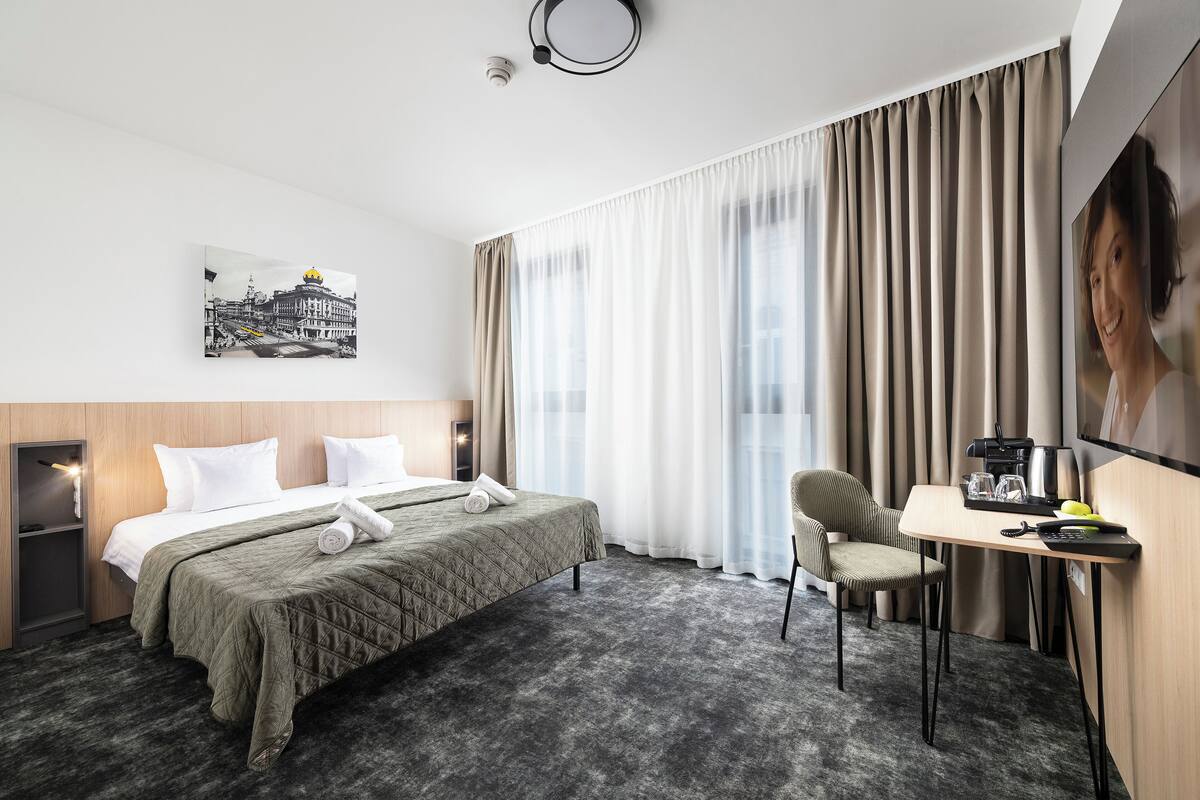 A hotel room with a bed, a desk, a chair, a TV, and a painting on the wall.