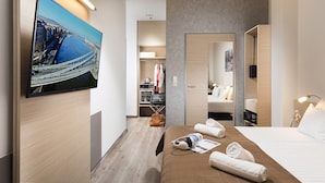 Quarto de hotel moderno com uma televisão de tela plana grande fixada na parede, cama bem arrumada com roupa de cama branca, piso de madeira e um closet espaçoso com roupas penduradas e uma mala.