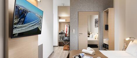 Ein modernes Hotelzimmer mit einem großen Flachbildfernseher an der Wand, einem ordentlich bezogenen Bett mit weißer Bettwäsche, Holzboden und einem begehbaren Kleiderschrank mit Kleidung und Koffer.