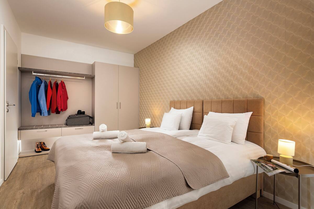 A neatly made bed with a beige bedspread, a bedside table with a lamp, and a wall with a geometric pattern.