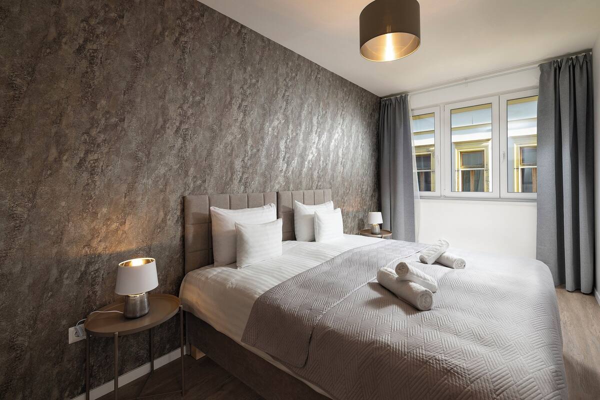 A modern hotel room with a large bed, a bedside table with a lamp, and a view of the city through the window.