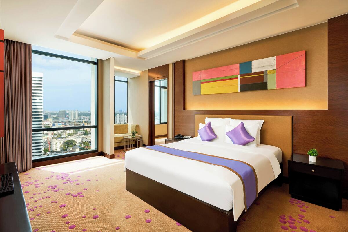A modern hotel room with a large bed, a city view, a desk, and a small plant on the nightstand.
