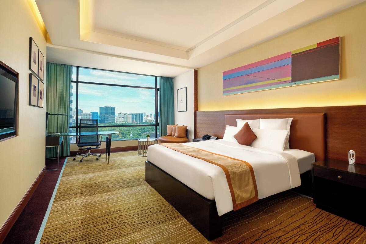A hotel room with a large bed, a desk, a chair, a TV, and a view of the city.