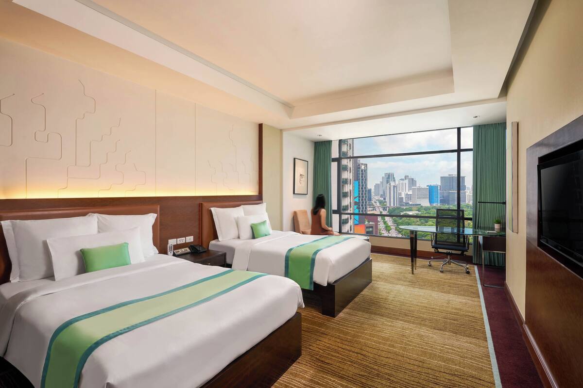 A hotel room with two beds, a large window with a city view, a desk, and a TV.